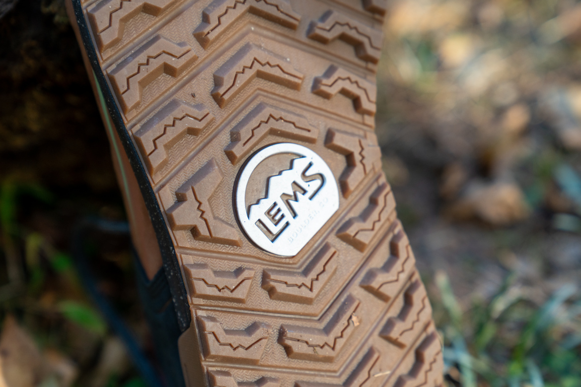 Close-up image of the textured sole of a shoe featuring a mountain design and the logo "LEMS." The outsole exhibits a rugged tread pattern, suitable for outdoor activities, with earthy tones.