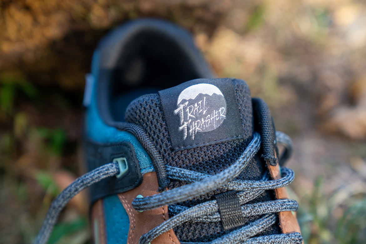 Close-up image of a sneaker featuring a "Trail Thrasher" logo on the tongue. The shoe has a combination of blue, brown, and gray materials, with textured fabric and laces prominently displayed. The background is slightly blurred, showcasing a natural outdoor setting with hints of grass and foliage.