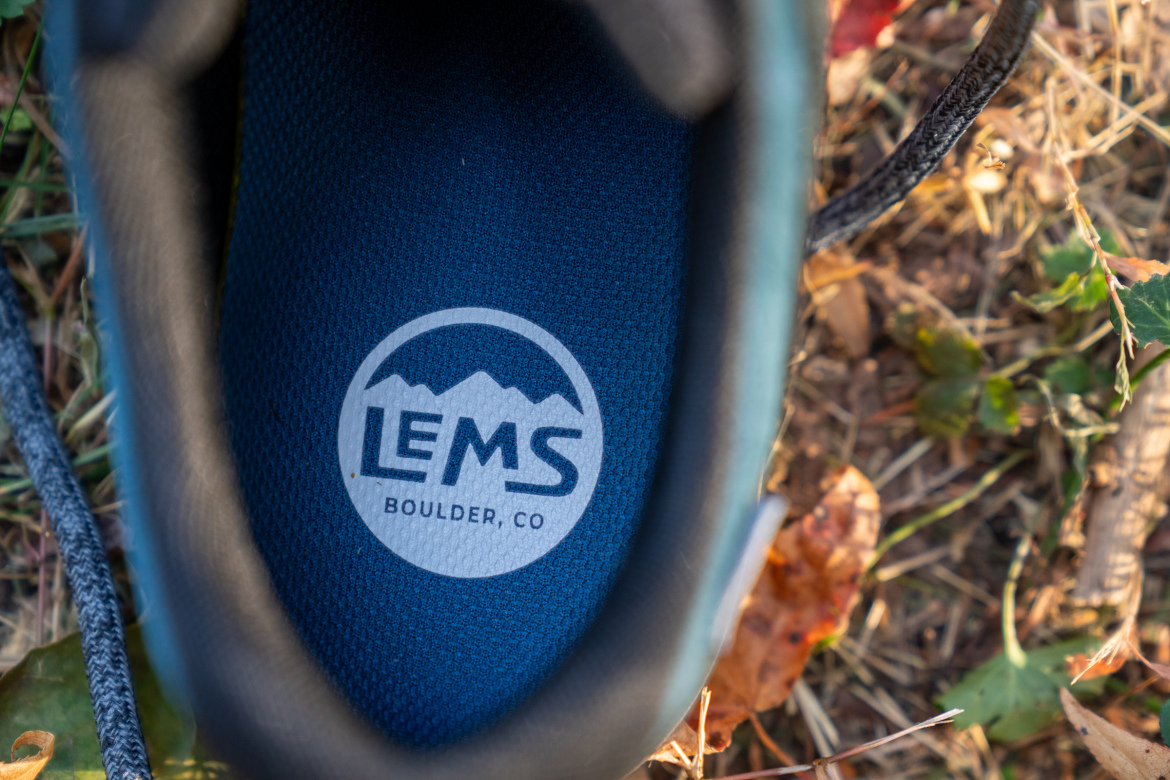 Close-up of the insole of a shoe featuring the Lems brand logo with the text "BOULDER, CO" underneath, set against a textured blue background. The shoe is positioned on natural ground with scattered leaves and grass.