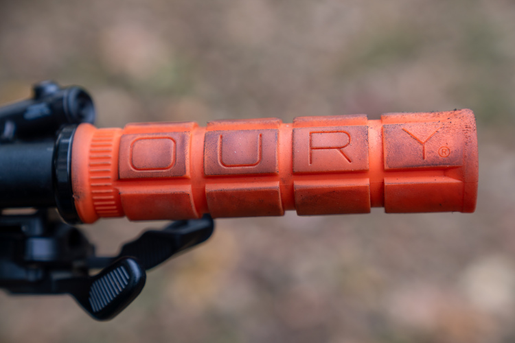 A close-up view of an orange rubber handle grip with the embossed letters "OURY" on it, mounted on a black bicycle handlebar. The grip appears slightly worn and dirty, suggesting usage in outdoor settings. The background is out of focus, highlighting the grip.