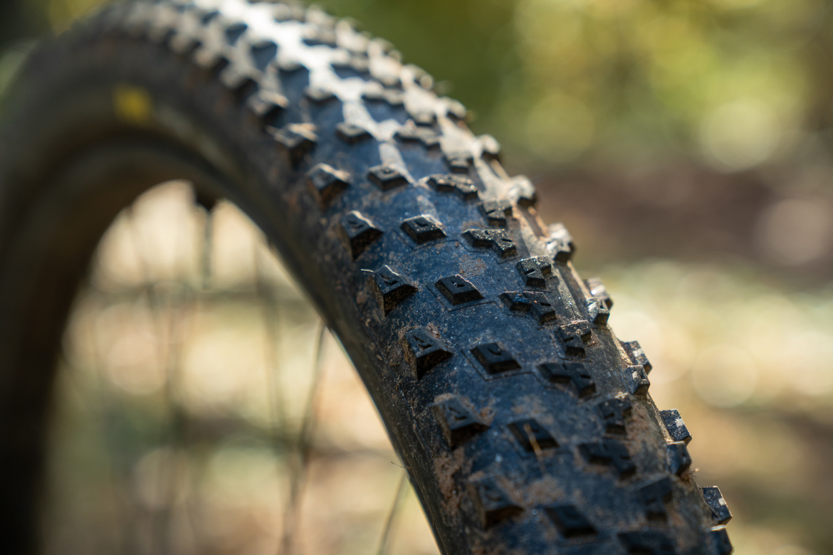 Close-up view of a mountain bike tire with a textured tread pattern, showing dirt and debris. The background is softly blurred, indicating an outdoor environment.