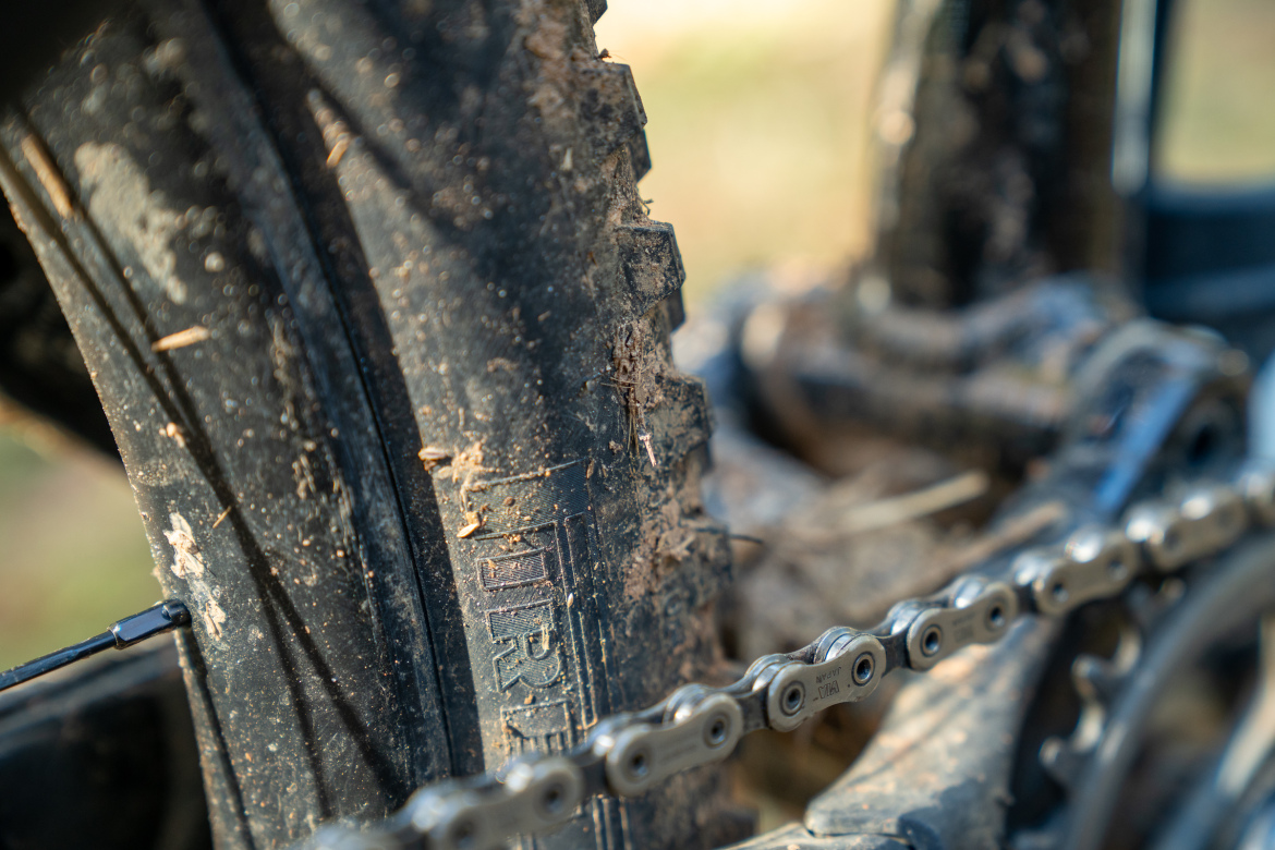 Close-up view of a muddy mountain bike tire and chain, showcasing the textured tread of the tire and the links of the chain against a blurred background of the bike frame and natural outdoor setting. The tire shows signs of dirt and debris, indicating off-road use.
