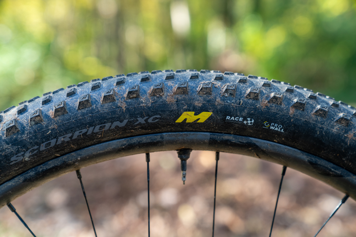 Close-up of a mountain bike tire featuring the tread pattern and branding of the Scorpion XC model. The tire shows signs of dirt, indicating recent use in outdoor conditions. In the background, a blurred natural setting with greenery suggests the tire is designed for trail riding.