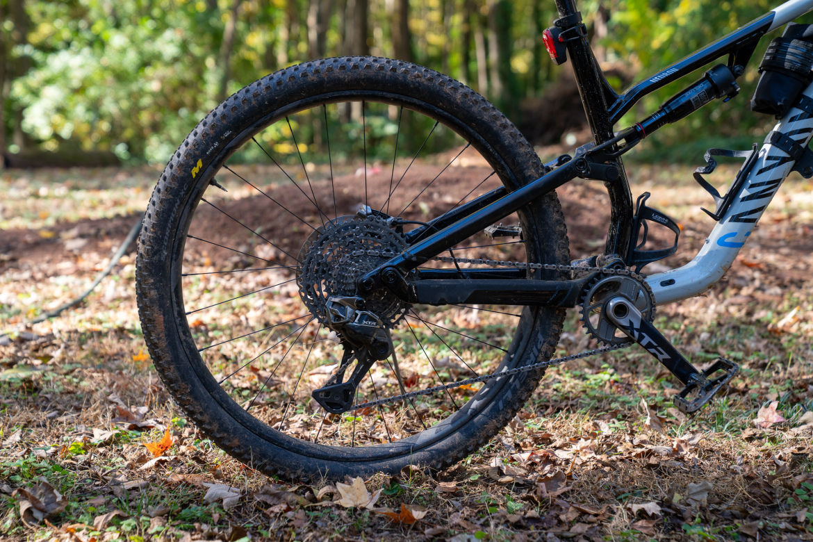A close-up view of the rear wheel and drivetrain of a mountain bike, featuring a muddy tire, gears, and chain. The background shows a natural setting with scattered leaves and greenery.