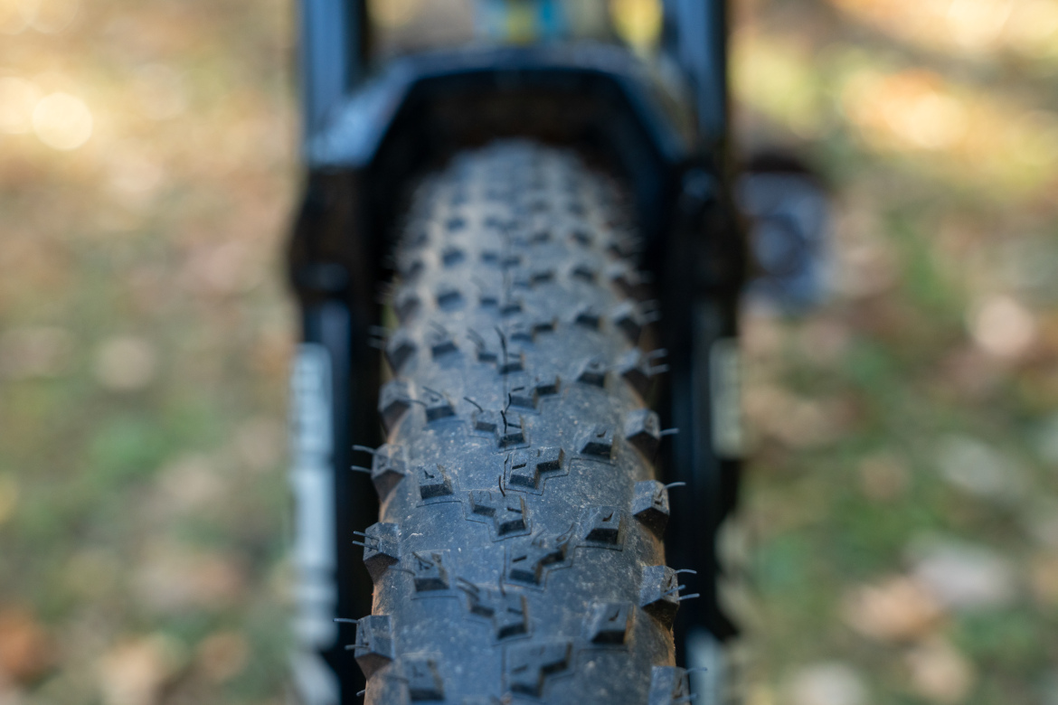 Close-up of a mountain bike tire tread, showcasing its textured surface with prominent, angled knobs designed for traction. The background features a blurred natural setting with fallen leaves, suggesting an outdoor trail environment.