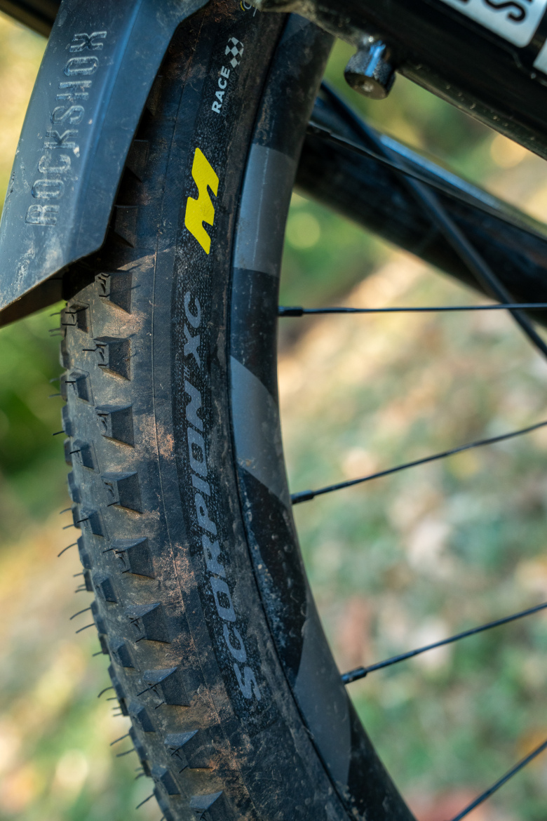 Close-up view of a bicycle tire featuring the brand name "Scorpion XC" and a prominent yellow "M" logo. The tire has a rugged tread pattern and is slightly dusted with dirt, indicating outdoor use. A portion of a bike's fork is visible in the image, along with blurred greenery in the background.