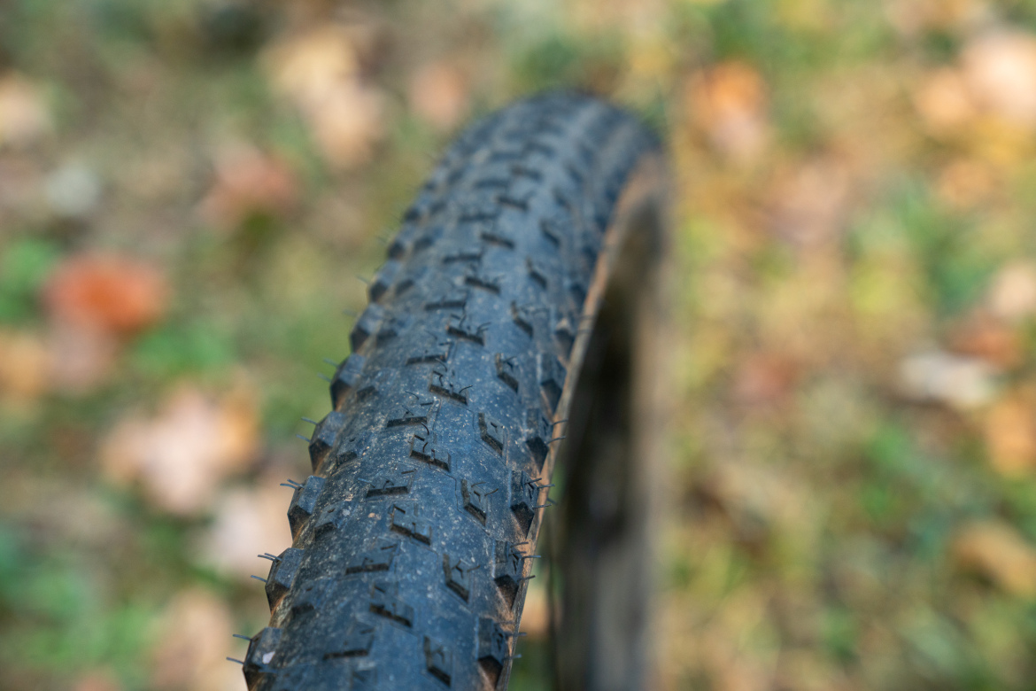 Close-up of a mountain bike tire with a textured tread pattern, set against a blurred backdrop of autumn leaves on the ground.