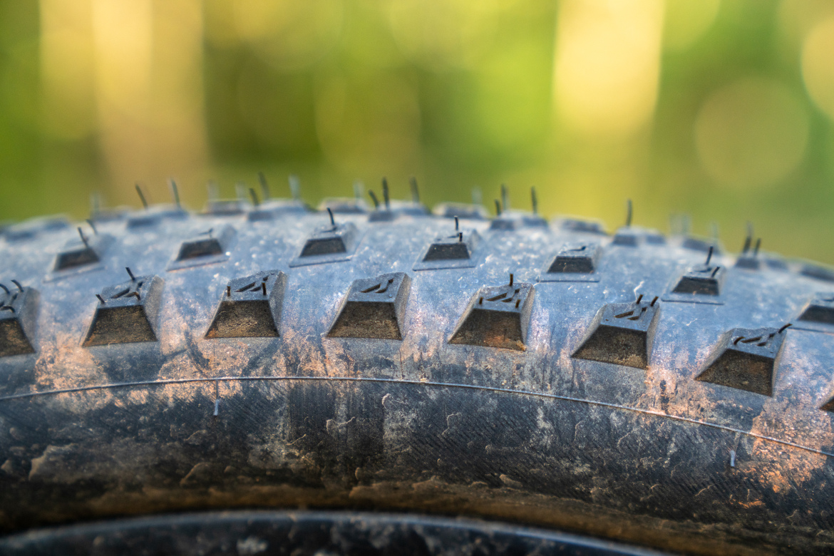 Close-up view of a mountain bike tire, showcasing its tread pattern with distinct angular protrusions and some dirt residue. The background features blurred greenery, suggesting an outdoor setting.