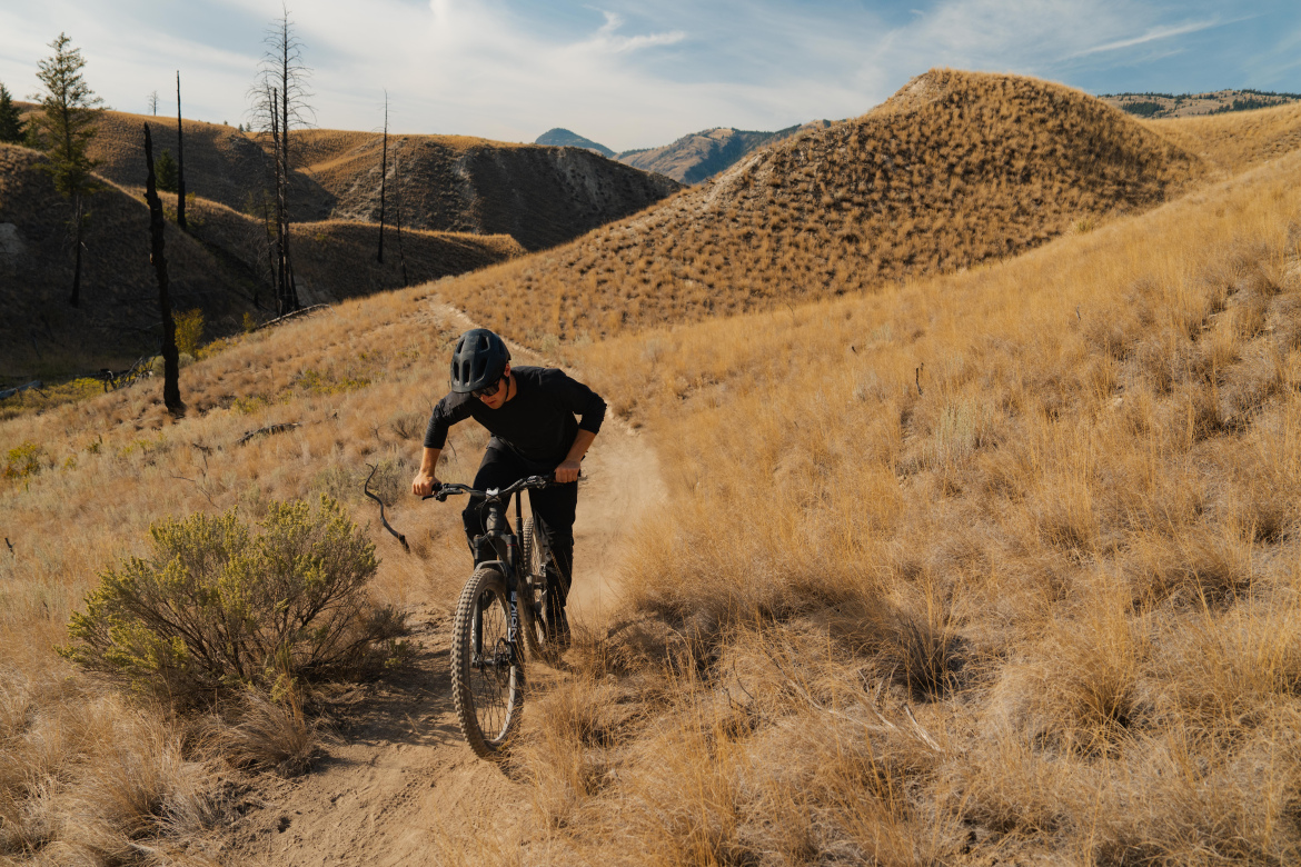 A mountain biker navigating a dirt trail through a vast landscape of golden grass and rolling hills. The cyclist, wearing a black helmet and attire, appears focused as they pedal uphill, surrounded by a natural setting with sparse vegetation and distant mountains under a clear blue sky.