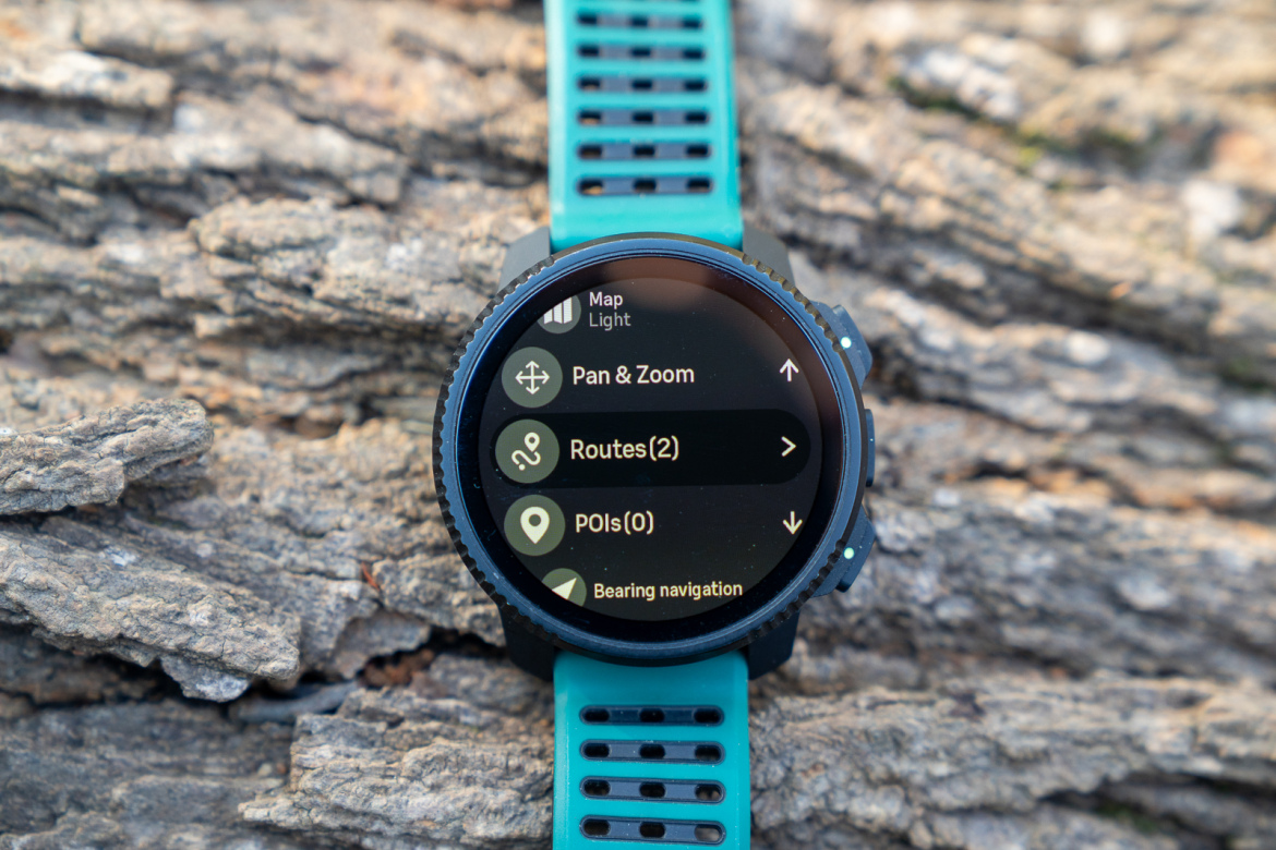A close-up of a smartwatch displaying a map interface, featuring options like "Pan & Zoom," "Routes," and "POIs," placed on a textured wooden surface. The watch has a black circular face with a turquoise strap.