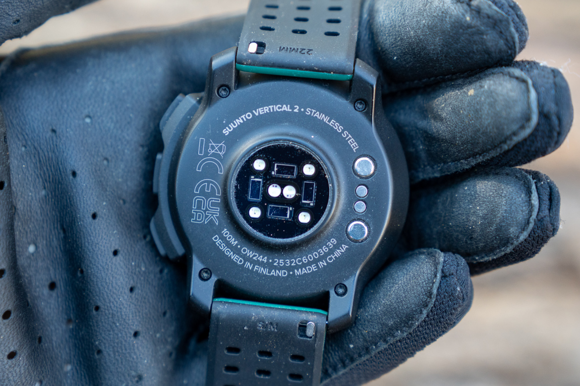 The back of a Suunto Vertical 2 watch with stainless steel detailing, held in a gloved hand. The watch features sensors and engraving detailing, including "DESIGNED IN FINLAND" and "MADE IN CHINA." The strap is black with ventilation holes.