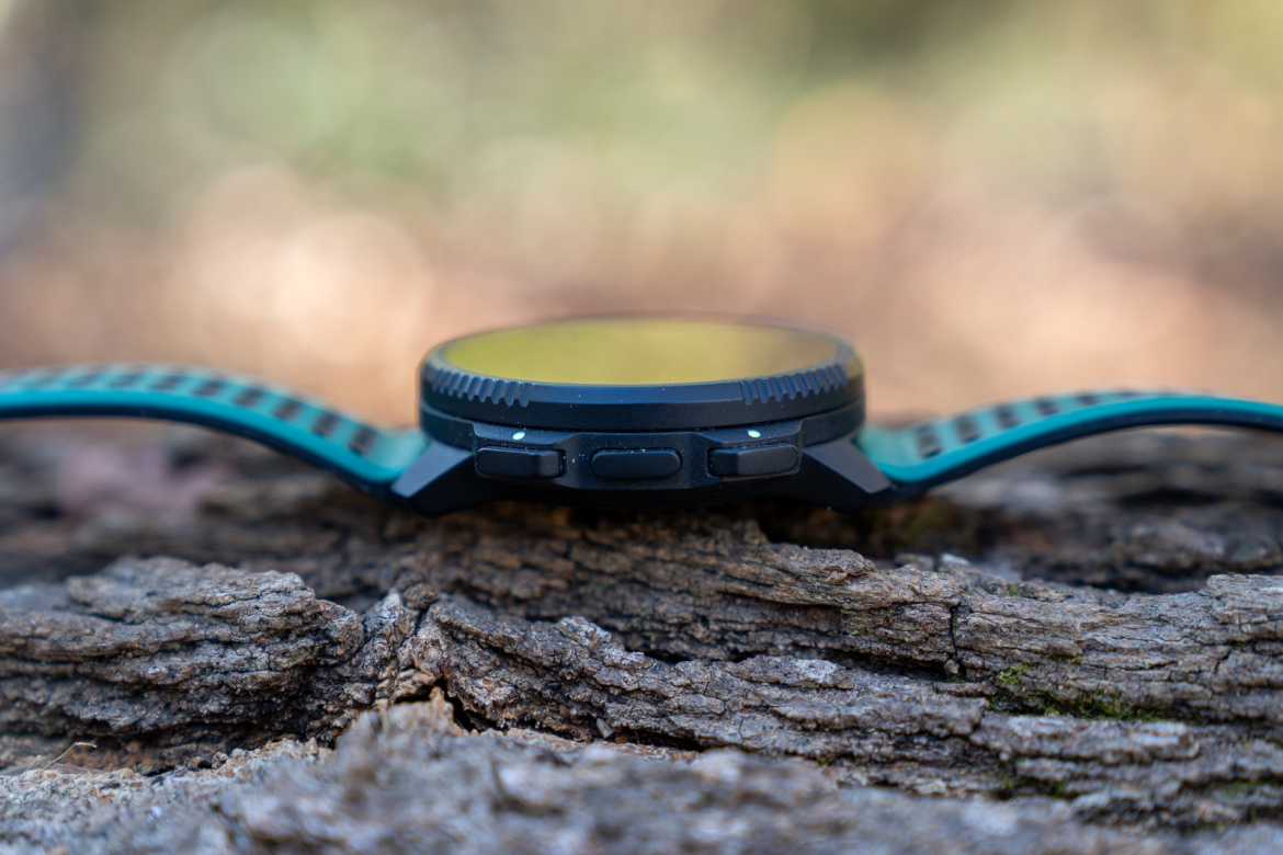 A close-up view of a black and teal sports watch resting on a wooden surface, showcasing its profile with buttons visible on the side. The background is soft-focused, highlighting the watch against a blurred natural setting.