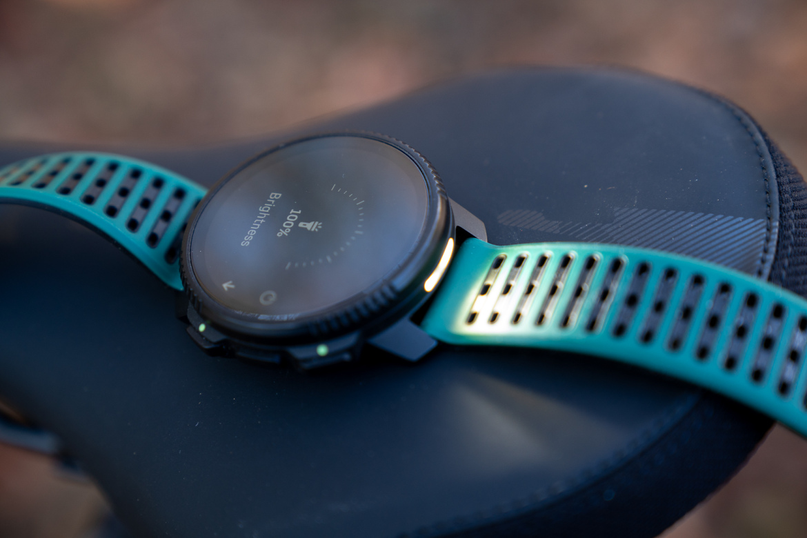 A close-up image of a smartwatch with a black face displaying a brightness setting of 100%, resting on a textured black surface, possibly a bike seat. The watch features a turquoise strap with ventilation holes.
