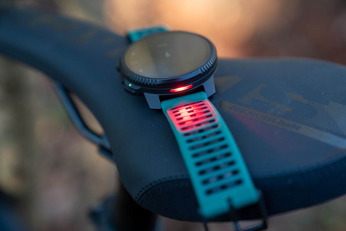 A close-up image of a fitness watch with a black face and teal strap, positioned on a bicycle saddle. The watch displays a red light on the back, indicating it may feature heart rate monitoring or notifications. The background is softly blurred, suggesting an outdoor setting.