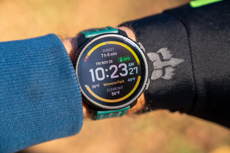 A close-up view of a smartwatch displaying the time as 10:23 AM on November 28, with additional information such as temperature and weather conditions. The wrist holding the watch is partially covered by a blue long-sleeve shirt, and another wrist is shown with a black sleeve featuring a logo. The background shows a blurred outdoor scene.