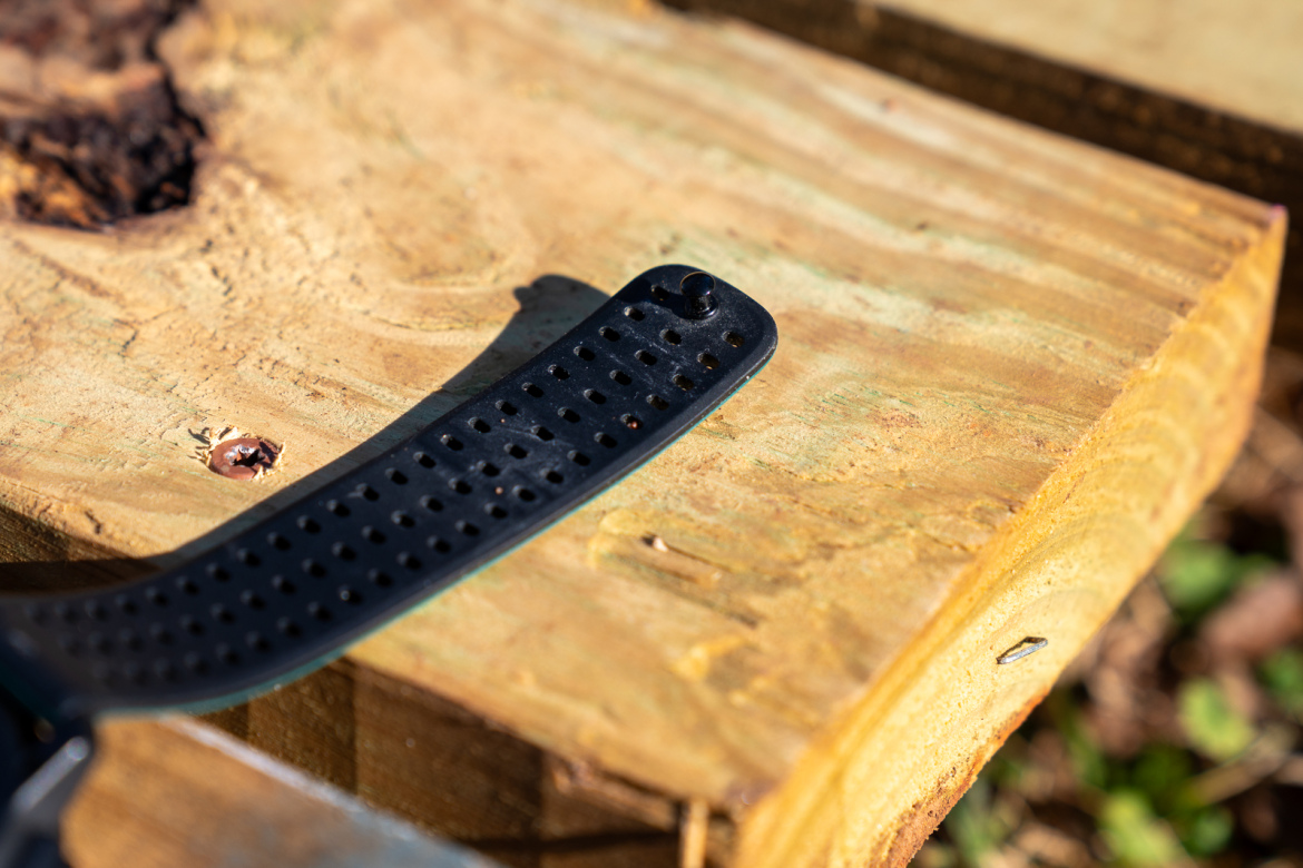 A close-up of a black rubber wristband placed on a wooden surface, featuring visible holes and a rounded end, with a small metal screw embedded in the wood nearby.