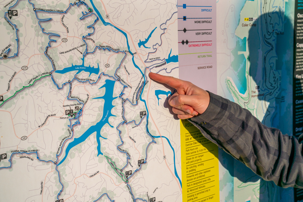 A person pointing at a detailed map showing the Back 40 trails in Bella Vista, Arkansas that highlights various trails and locations in a park, with marked paths indicating different difficulty levels. The map includes multiple lakes and forests, along with information on parking and regulations.