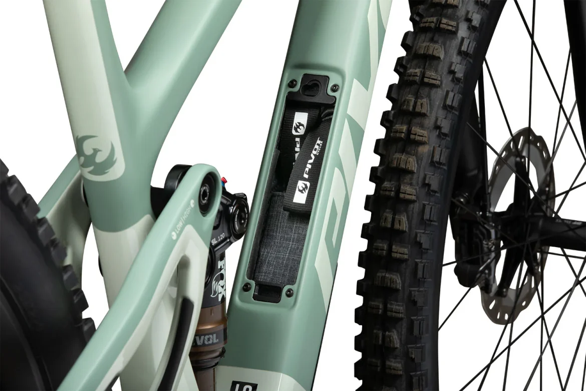 Close-up of a mountain bike frame featuring a mint green color scheme, showcasing the rear shock and a storage compartment integrated into the frame. The image highlights the bike's intricate design and tire tread.