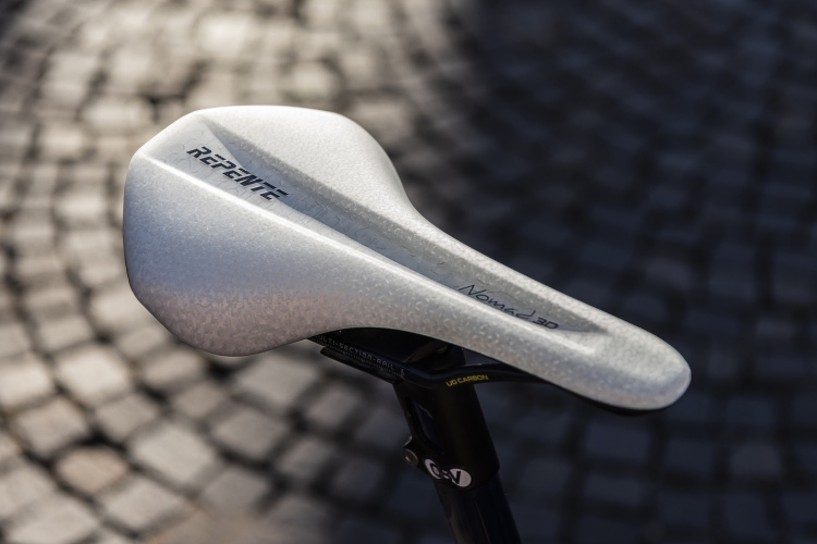 A close-up view of a white bicycle saddle featuring a sleek design with branding that reads "Repente Nomad 3D," positioned against a cobblestone surface. The saddle has a textured finish, emphasizing its modern aesthetic.