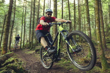 Two mountain bikers navigating a wooded trail, one rider performing a jump while wearing a red shirt and sunglasses. The forest is lush with green foliage and tall trees, providing a natural backdrop for the adventurous scene. Trowbridge Forest / Shuniah Mines mountain bike trail.