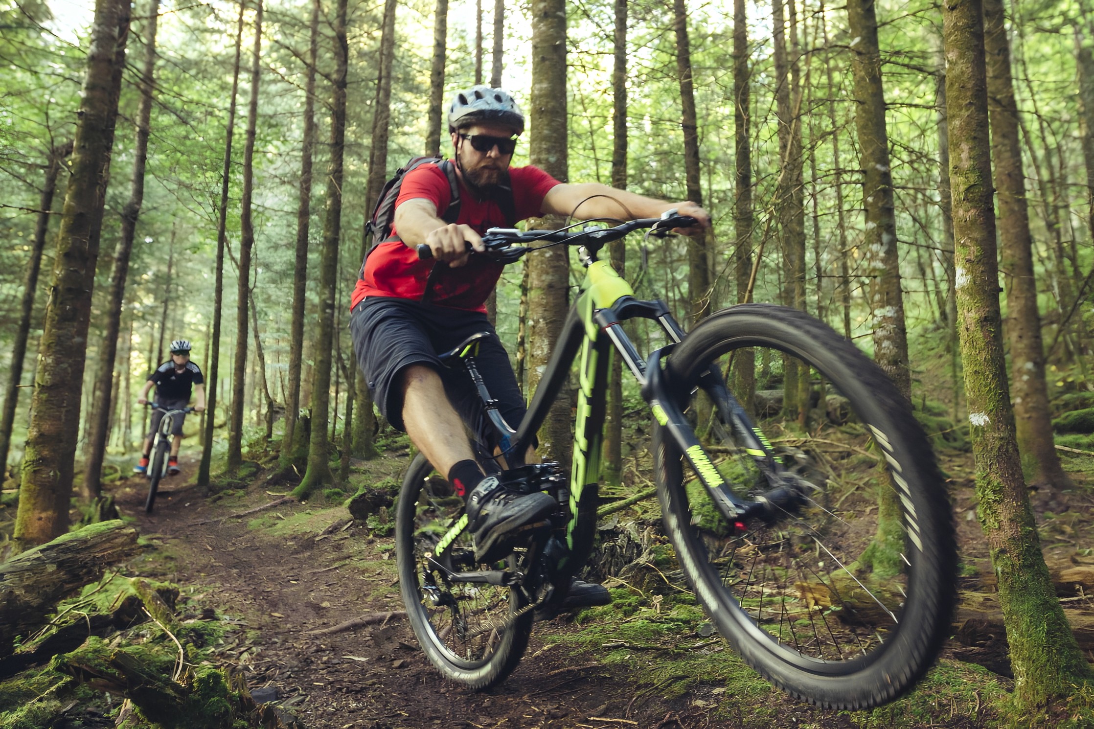 Two mountain bikers navigating a wooded trail, one rider performing a jump while wearing a red shirt and sunglasses. The forest is lush with green foliage and tall trees, providing a natural backdrop for the adventurous scene. Trowbridge Forest / Shuniah Mines mountain bike trail.
