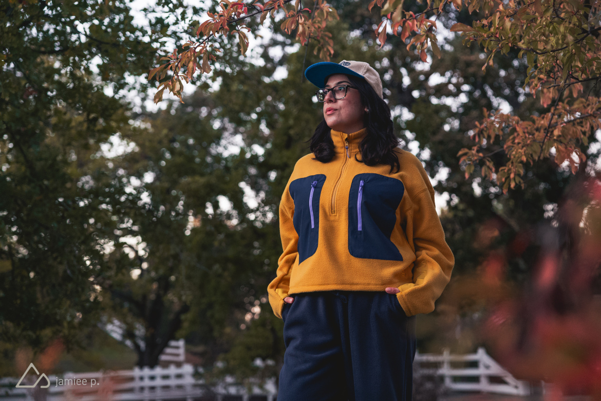 A person wearing a bright yellow fleece pullover with navy blue accents stands outdoors among autumn foliage. They have dark, wavy hair, glasses, and are wearing a beige cap. The background features trees with green leaves transitioning to autumn colors.