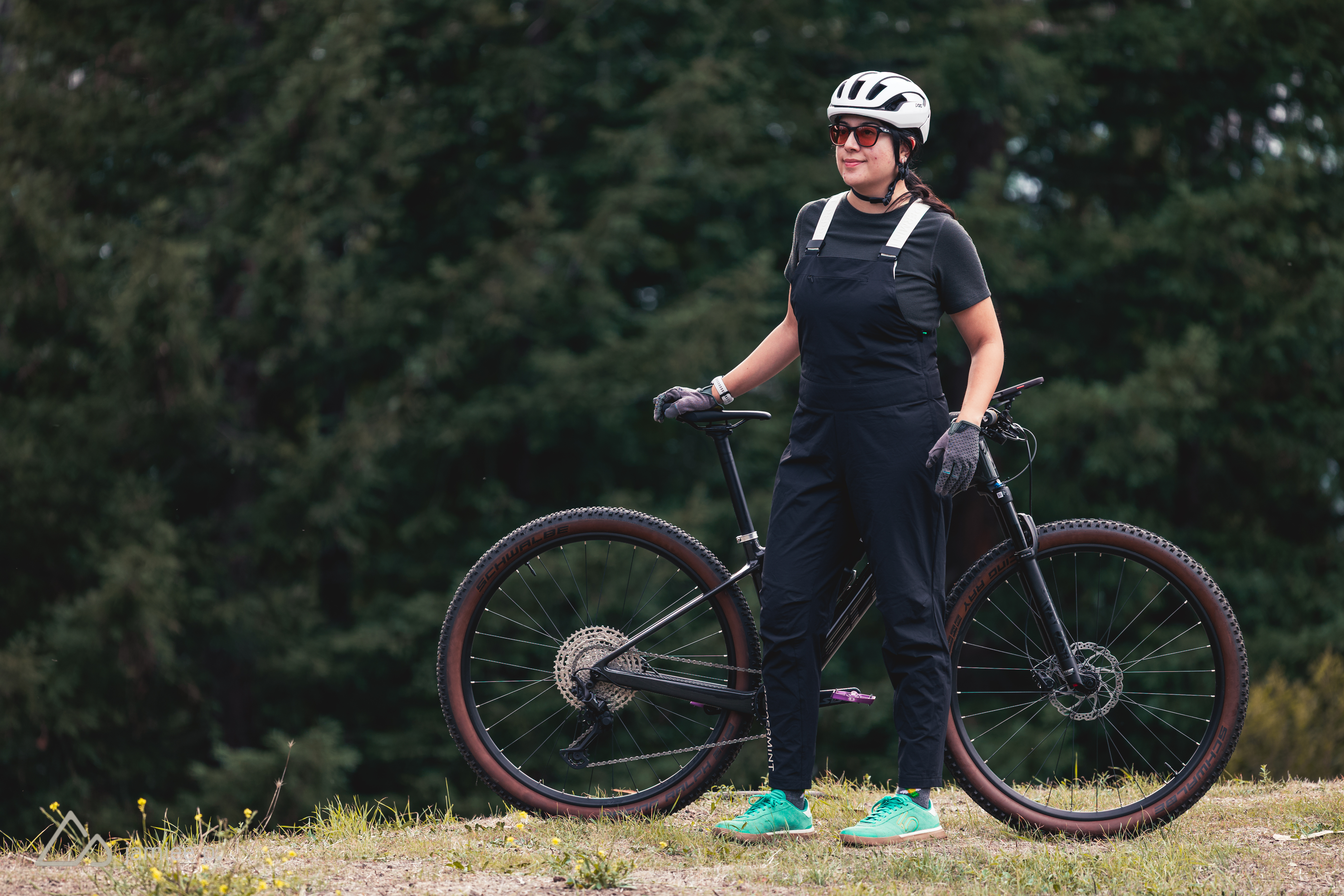 A woman in a black cycling outfit and helmet stands beside her mountain bike on a grassy hill, surrounded by trees. She wears sunglasses and gloves, exuding a sporty and adventurous vibe.