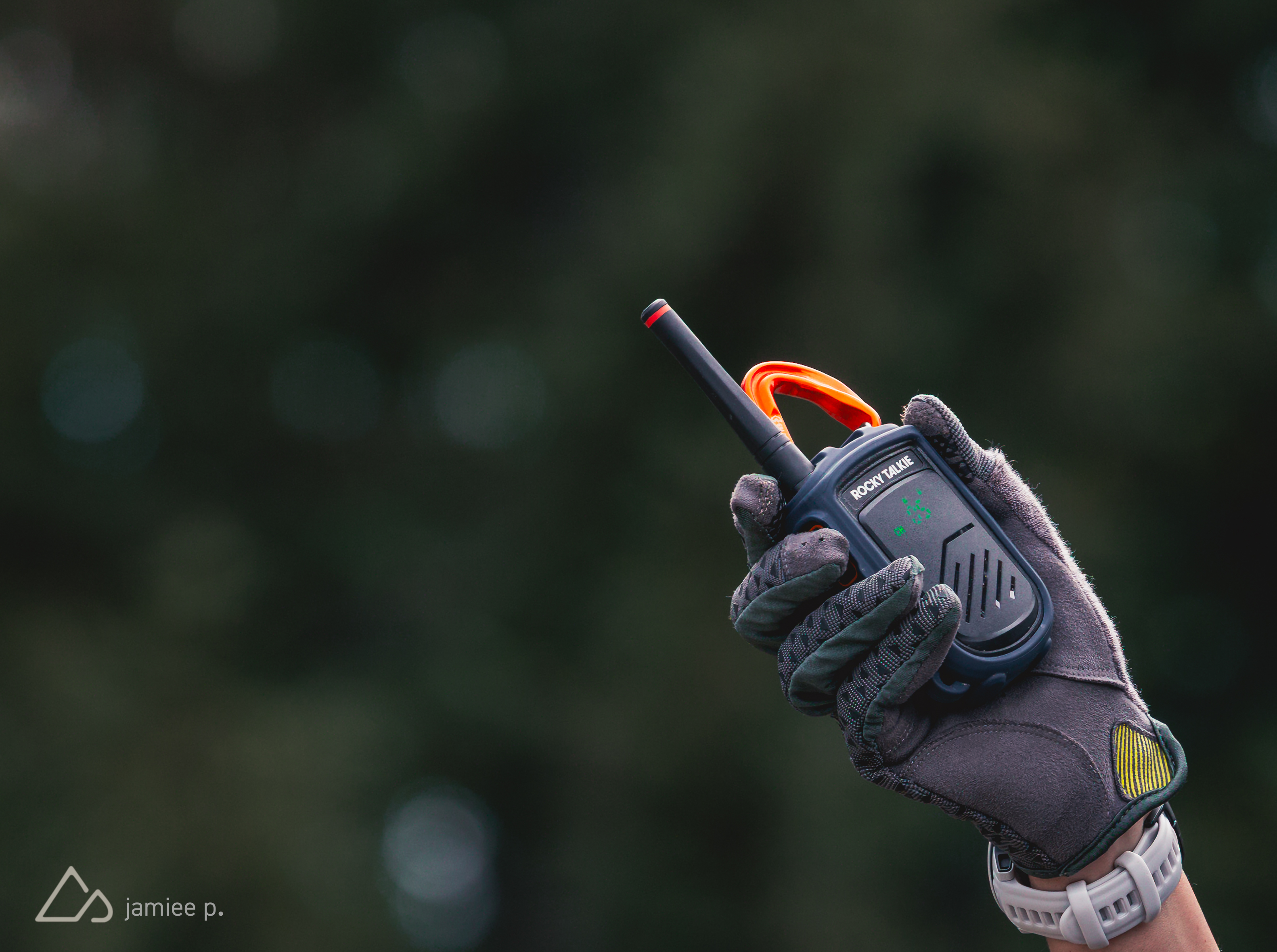 A close-up image of a hand wearing a gray glove, holding a black two-way radio with an orange antenna. The background is softly blurred, suggesting an outdoor setting. The device displays green indicators on its screen.