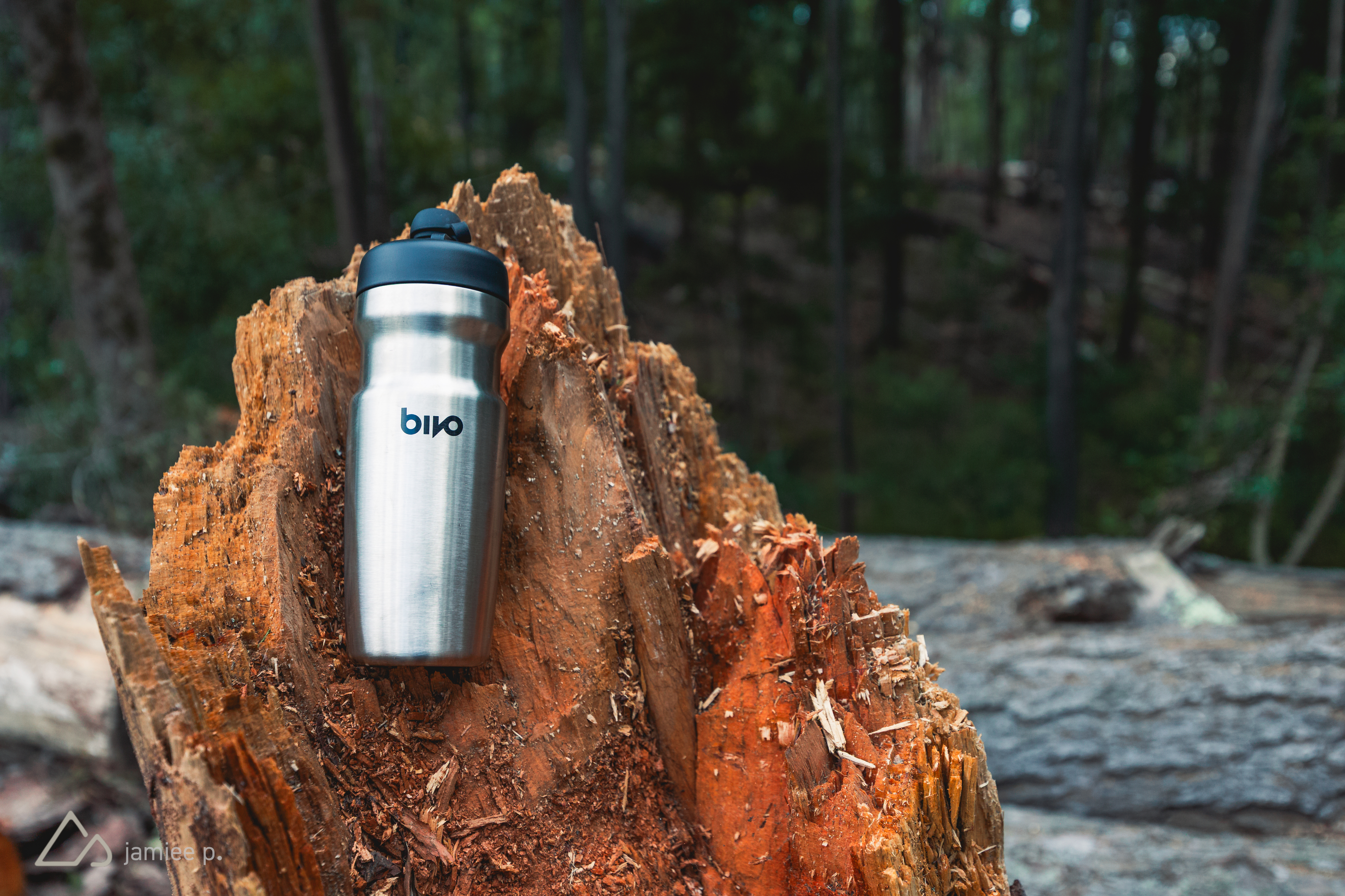 A stainless steel water bottle resting on a textured tree stump, surrounded by a lush green forest. The bottle features a black lid and the logo "bivo" clearly visible on its surface.