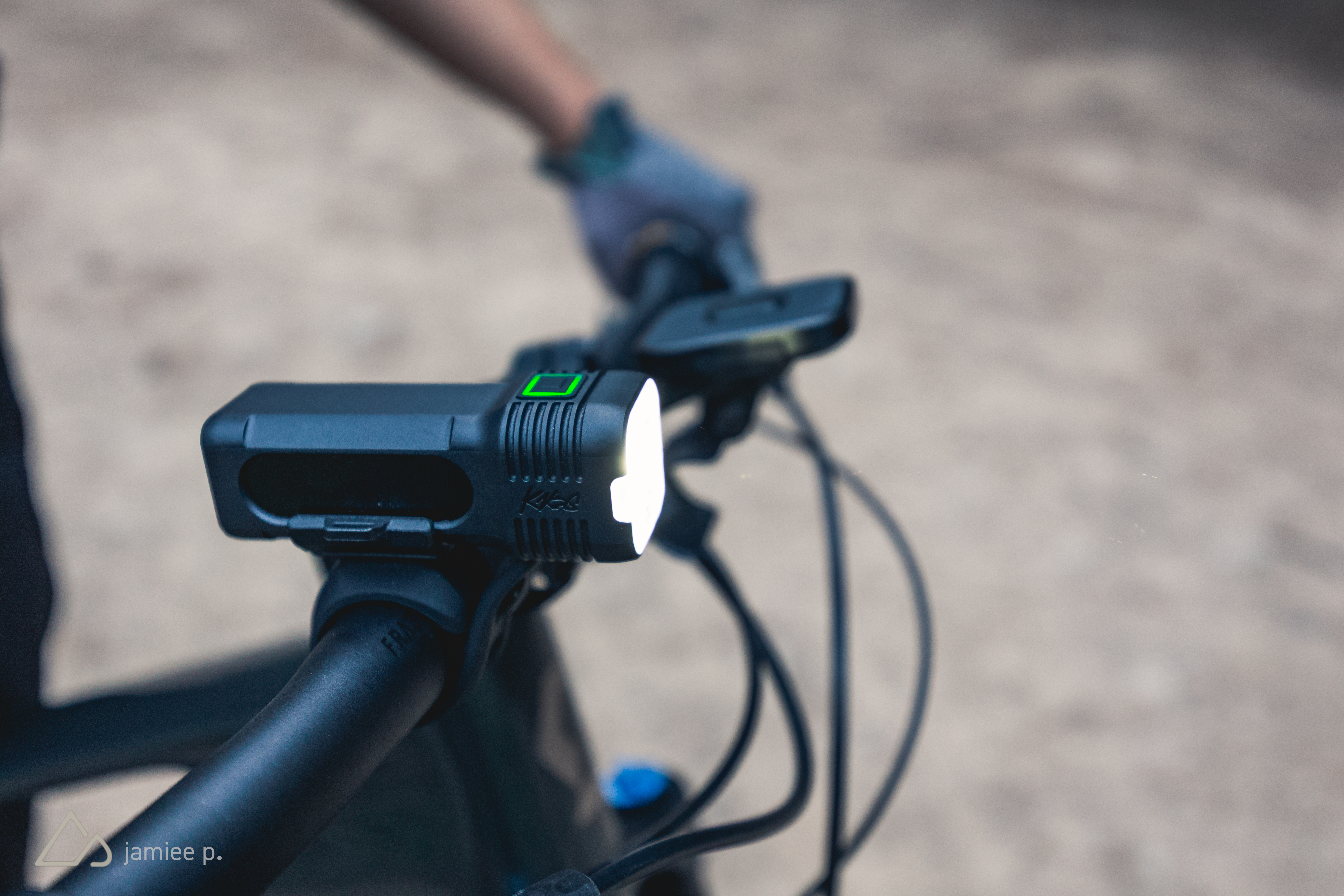 A close-up view of a bicycle's handlebar featuring a mounted LED light. The light is black with a white front and a green indicator light. In the background, a person's gloved hand is gripping the handlebar, and the ground is visible, suggesting an outdoor setting.