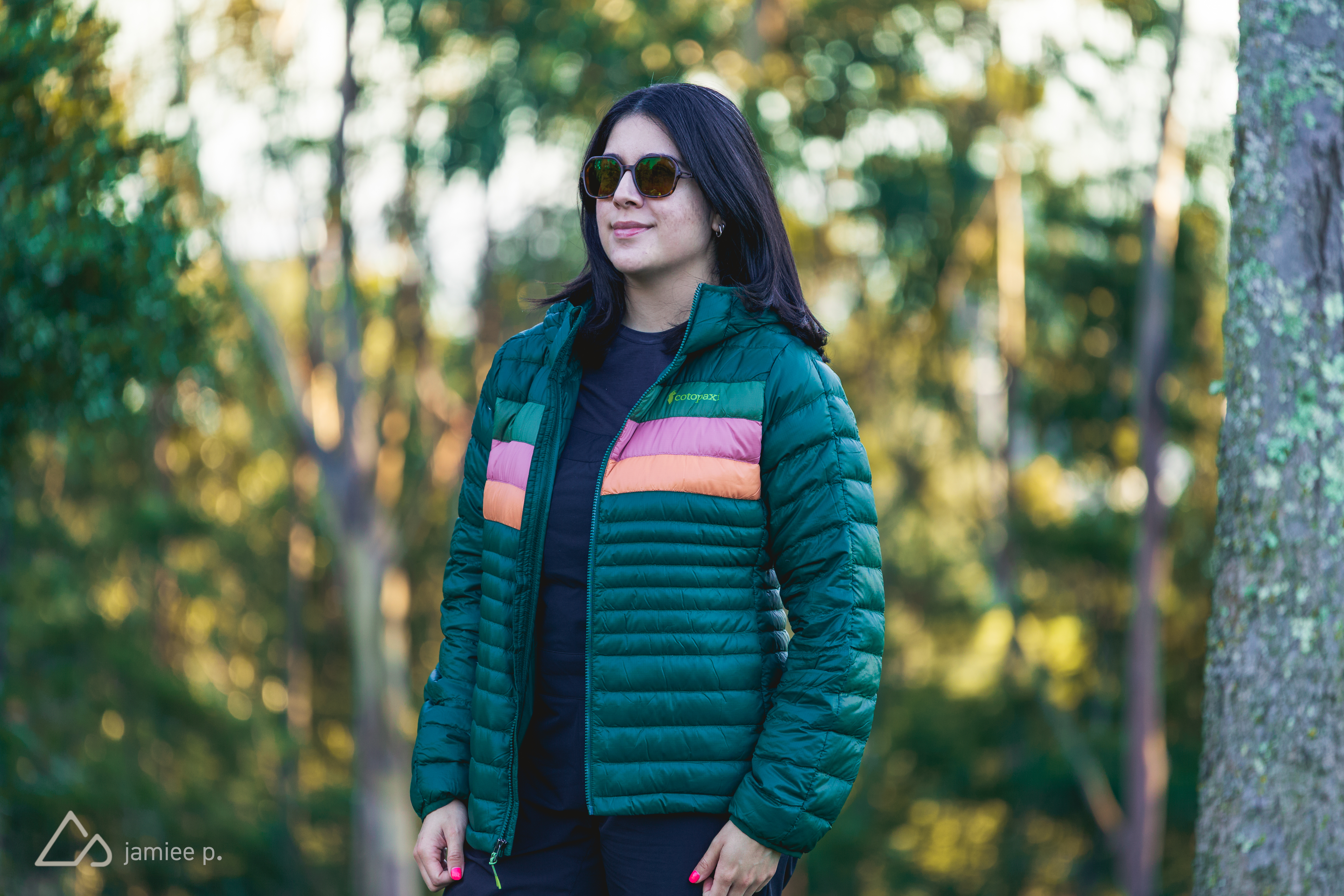 A person stands outdoors in a forested area wearing a green puffer jacket with colorful horizontal stripes, sunglasses, and a black shirt. The background features blurred trees and soft natural light, suggesting a pleasant day.
