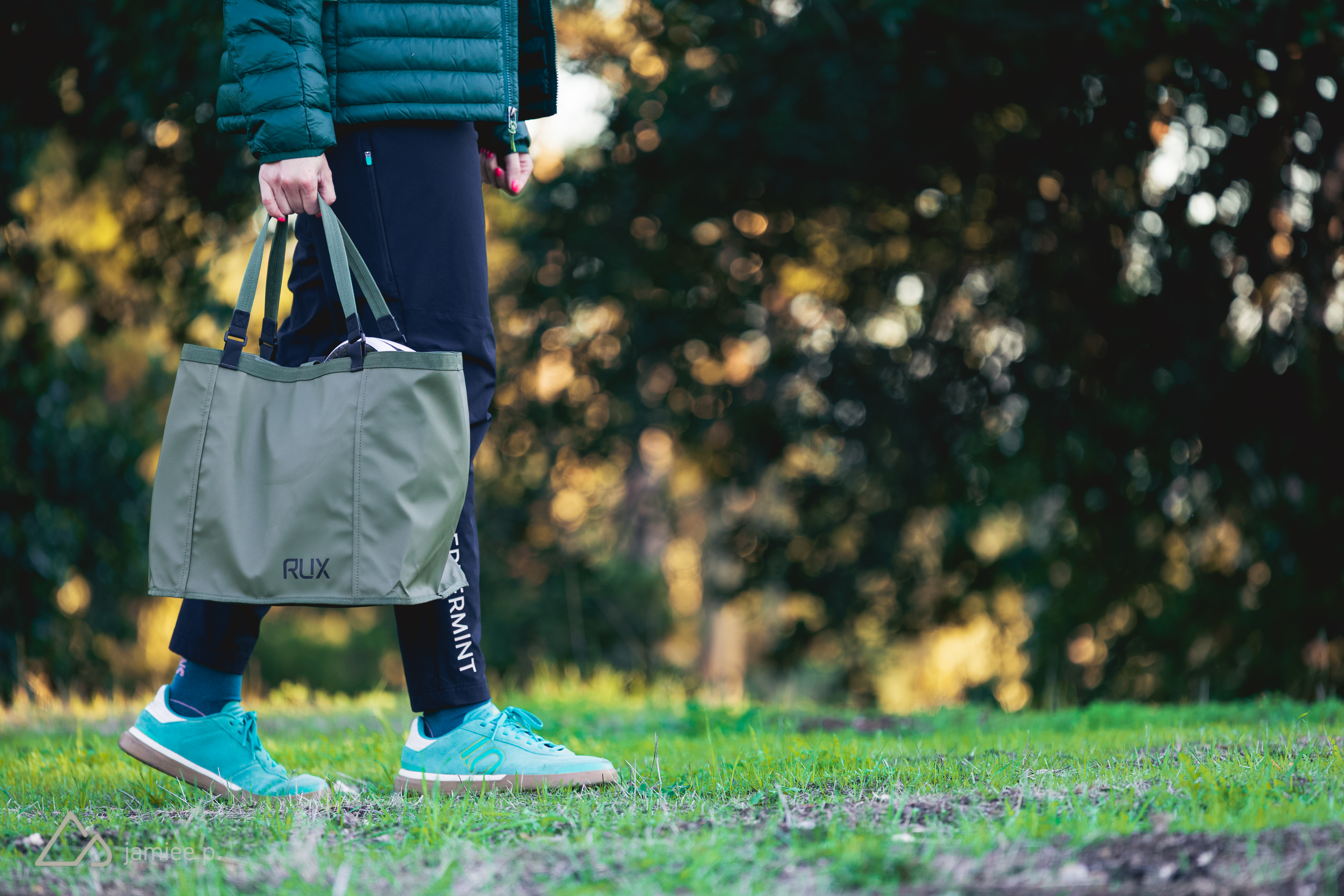 A person wearing a green puffer jacket and black pants is walking on grass, carrying a light green tote bag. They are wearing bright blue athletic shoes, and the background features blurred greenery, suggesting an outdoor setting.