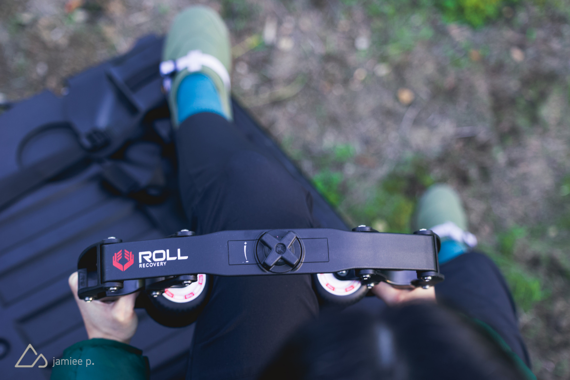 A close-up view of a person holding a Roll Recovery massage tool, featuring a black strap with wheels and red logo, while seated outdoors. The person's legs are partially visible, wearing green shoes and blue socks. The background shows a natural setting with grass and leaves.