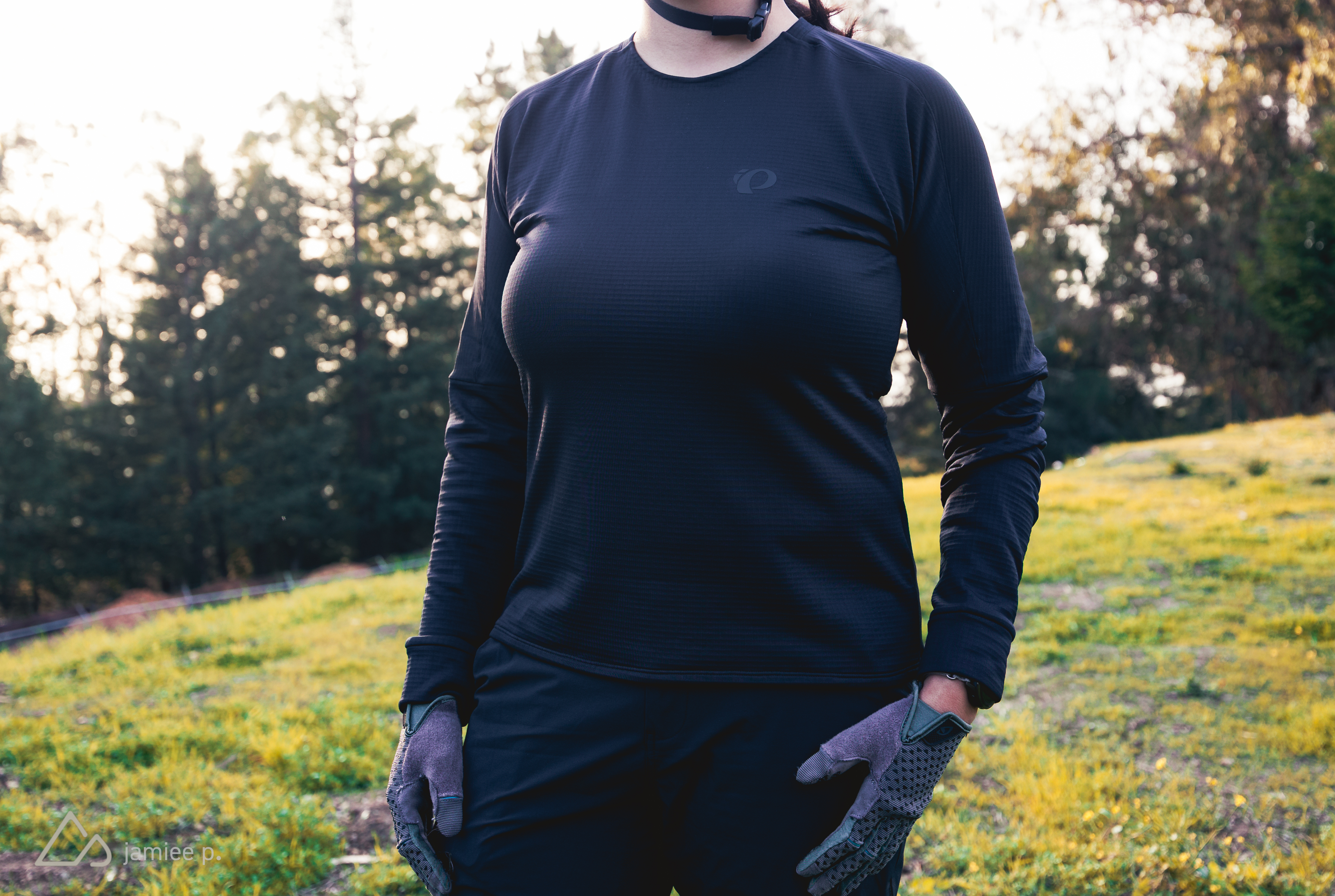 A person wearing a long-sleeved black athletic shirt and gloves stands outdoors in a grassy area, with trees in the background. The scene is well-lit, suggesting a sunny day.