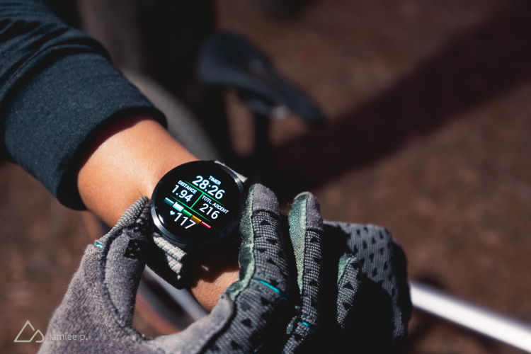 A close-up view of a hand wearing a black watch displaying fitness metrics, including timer, distance, total ascent, and heart rate. The hand is partially gloved, and the background features soft, natural lighting, suggesting an outdoor activity.