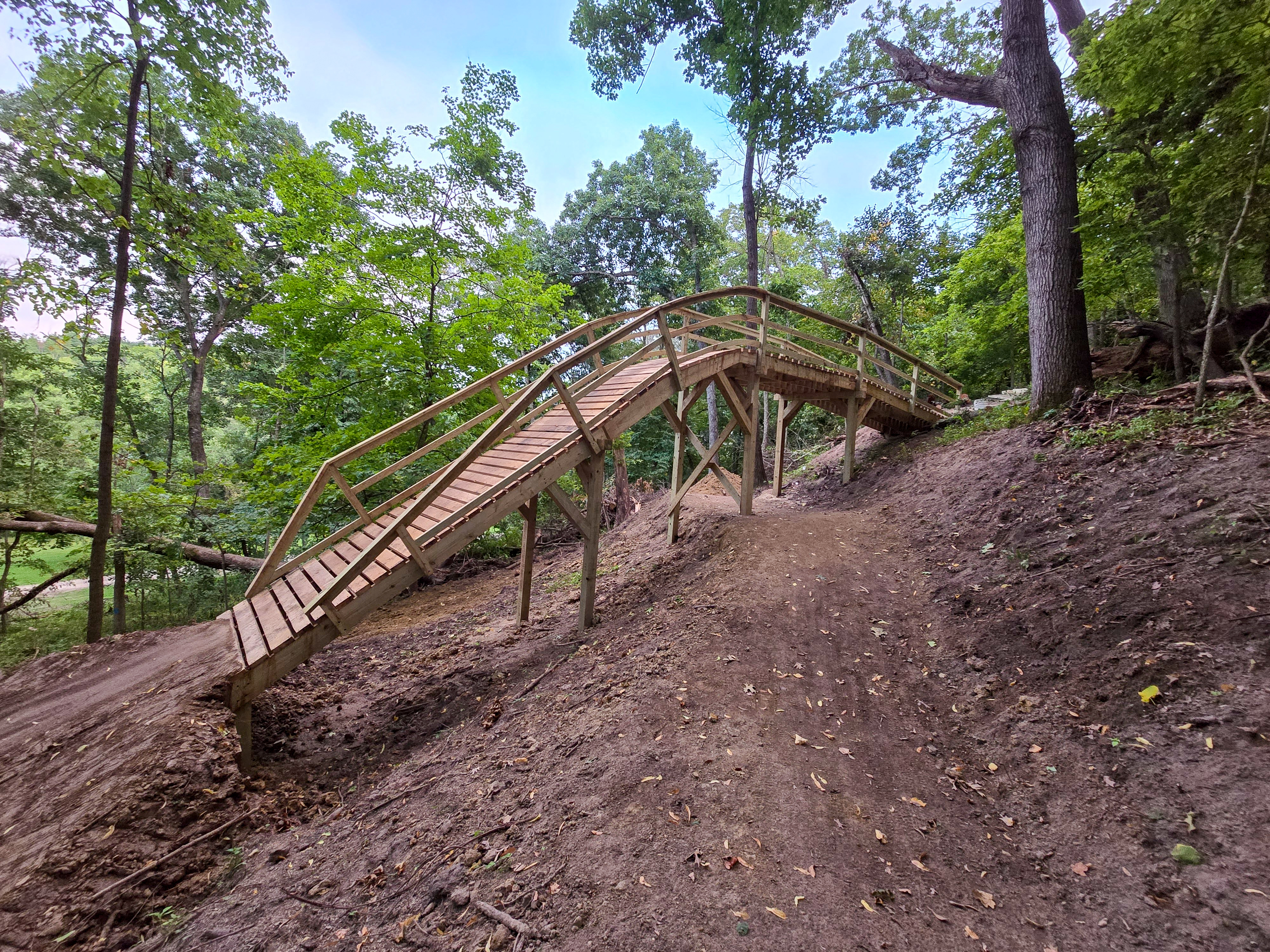 Wooden bridge built on a slope surrounded by lush green trees, leading over a dirt path in a forested area. Pinicon Ridge Park mountain bike trail.