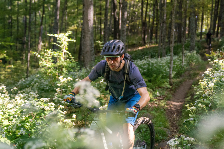 A person riding a mountain bike on a narrow trail through a lush green forest, surrounded by wildflowers and trees. The rider is wearing a helmet, sunglasses, and biking gear, appearing focused as they navigate the terrain.