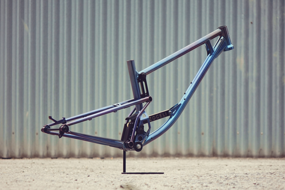 A blue and black mountain bike frame displayed upright against a textured, corrugated metal background. The frame features modern geometric design elements and is propped on a small stand.