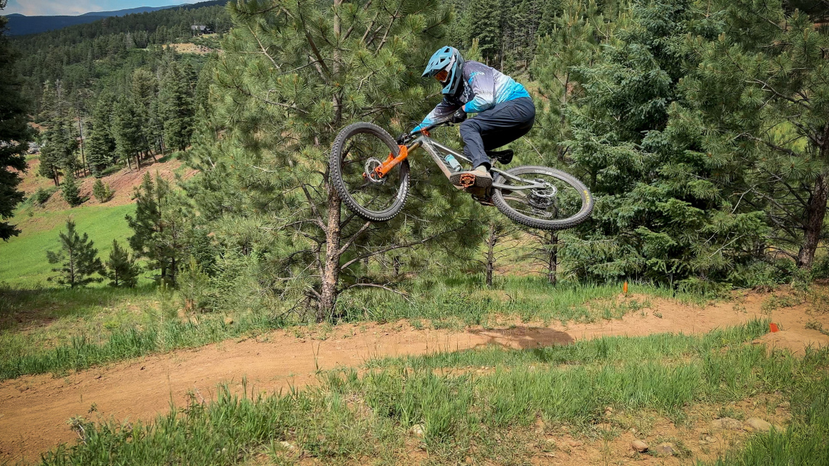 A mountain biker performing a jump on a dirt trail surrounded by pine trees, showcasing an action-packed moment with the bike's front wheel elevated off the ground. The rider is dressed in a blue helmet and jersey, highlighting the outdoor, adventurous atmosphere.