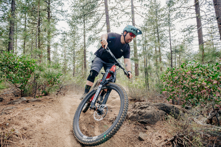 A mountain biker navigating a dirt trail in a forest, leaning into a turn with a trail of dust behind the bike, surrounded by tall trees and green bushes. The rider is wearing a helmet and protective gear.