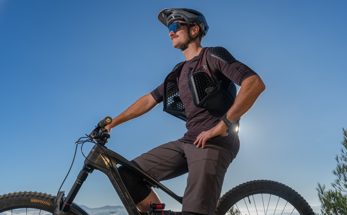 A mountain biker posing confidently on a bike, wearing a helmet and protective gear, against a clear blue sky. Trees and mountains are visible in the background, highlighting an outdoor adventure theme.