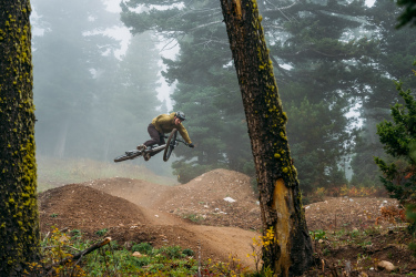 A mountain biker performs a jump over a dirt ramp in a foggy forest setting. The rider is wearing a helmet and protective gear, surrounded by tall trees and overgrown vegetation. The scene captures the action and thrill of mountain biking in a natural outdoor environment. Gnomadic mountain bike trail.