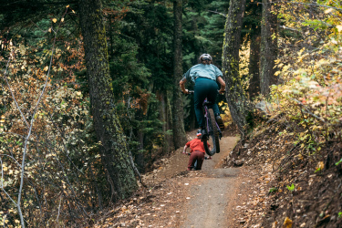 A person riding a mountain bike jumps off a small ramp on a dirt trail surrounded by tall trees and autumn foliage, while a child in a red outfit is seen biking close behind. Gnomadic mountain bike trail.