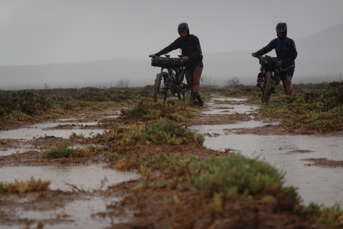 Two cyclists push their bikes along a muddy path in heavy rain. The landscape is a mix of wet soil and sparse vegetation, with gray clouds dominating the sky, creating a moody atmosphere.