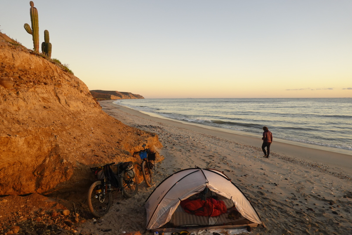 A scenic beach at sunset, featuring a tent set up on the sandy shore, with two bicycles parked nearby. A person in a red jacket strolls along the water's edge, while a rocky cliff with a cactus is visible in the background. The waves gently lap at the shore under a gradient sky transitioning from orange to blue.