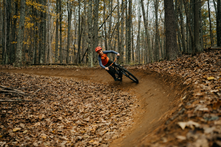 A mountain biker in an orange helmet and athletic gear leans into a sharp turn on a dirt trail surrounded by trees with autumn leaves scattered on the ground. The bike tires create a slight spray of dirt as the rider navigates the curve.