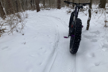 A fat-tire bike positioned on a snowy trail in a forest, with snow-covered trees in the background. The bike's rear wheel shows a tread pattern, and animal tracks can be seen in the fresh snow on the ground. Beach Ridge Singletrack mountain bike trail.