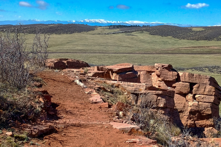 A scenic view of a rocky cliff in the foreground with a dirt path winding along it, surrounded by sparse vegetation. In the background, rolling green hills transition to snow-capped mountains under a bright blue sky with a few clouds.