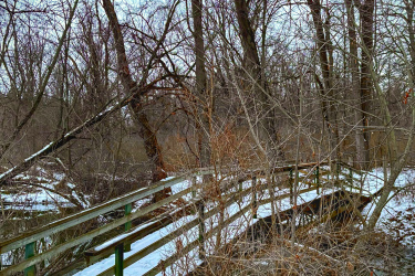 A snow-covered wooden bridge crossing a small stream, surrounded by bare trees and shrubs in a winter setting. The sky is overcast, creating a serene atmosphere in the natural landscape. Rotary Memorial Trail mountain bike trail.