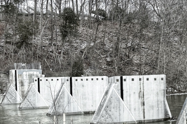 A concrete structure with triangular walls stands in a river, surrounded by a snowy landscape and bare trees. The scene is monochromatic, featuring muted tones of gray and white, with visible snow on the ground and branches. The backdrop includes a forested hill, adding depth to the composition. Springbank park mountain bike trail.